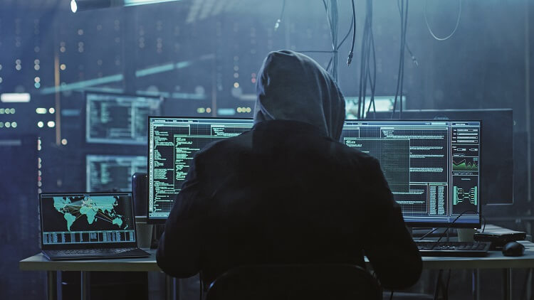 Hooded hacker at multiple computer screens committing identity theft